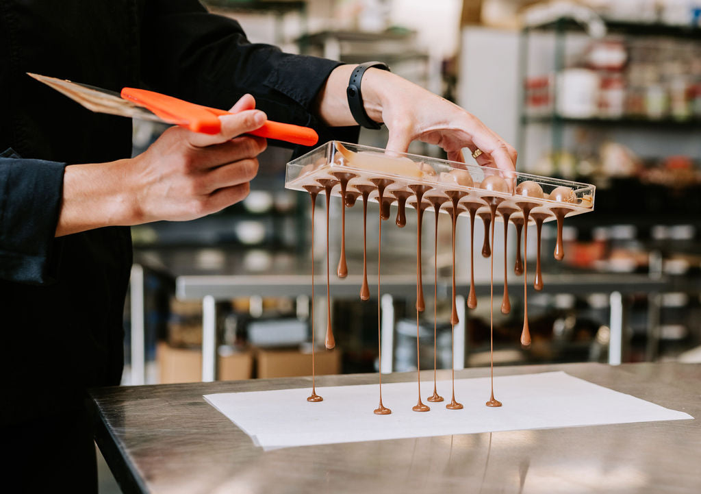 Chocolatier using a spatula and holding a chocolate mold that drips milk chocolate photo taken by Meaghan Baxter Photography